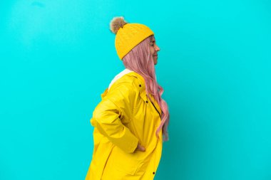 Pembe saçlı, yağmura dayanıklı bir ceket giyen, mavi arka planda izole edilmiş, gayret gösterdiği için sırt ağrısı çeken genç bir kadın.