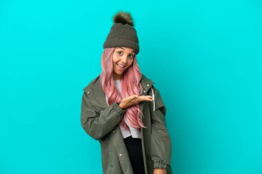 Pembe saçlı, yağmura dayanıklı bir ceket giyen genç bir kadın mavi arka planda izole edilmiş bir fikir sunuyor ve gülümsüyor.