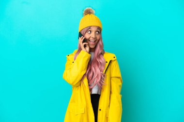 Pembe saçlı, yağmura dayanıklı bir ceket giyen genç bir kadın mavi arka planda izole edilmiş cep telefonuyla sohbet ediyor.