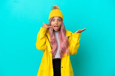 Pembe saçlı, yağmura dayanıklı bir ceket giyen genç bir kadın mavi arka planda izole edilmiş telefon jestleri yapıyor ve kuşkulanıyor.