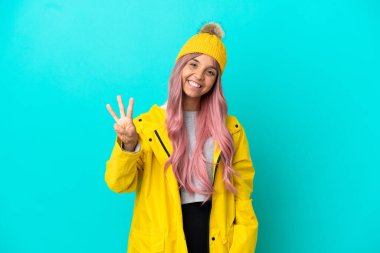 Pembe saçlı, yağmura dayanıklı bir ceket giyen genç bir kadın mavi arka planda izole edilmiş mutlu ve parmaklarıyla üçe kadar sayıyor.