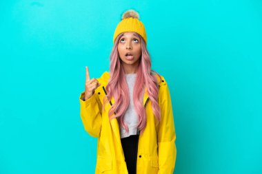 Pembe saçlı, yağmura dayanıklı bir ceket giyen genç bir kadın mavi arka planda izole edilmiş ve şaşırmış.