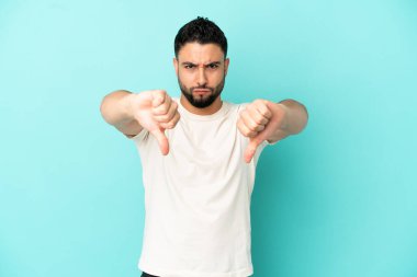 Mavi arka planda izole edilmiş genç bir Arap, baş parmağını iki eliyle aşağı gösteriyor.