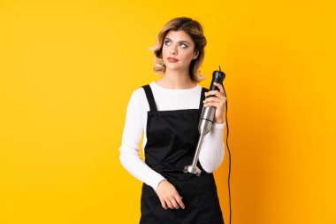 Sarı arka planda izole edilmiş el blenderi kullanan kız şüpheli hareketler yapıyor.