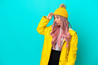 Pembe saçlı, yağmura dayanıklı bir ceket giyen genç bir kadın. Mavi arka planda yalıtılmış, sürpriz bir ifadeyle yan yana bakarken.
