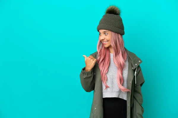 粉红头发的年轻女人穿着防雨外套 蓝色背景隔离 指向侧面来展示一种产品 — 图库照片