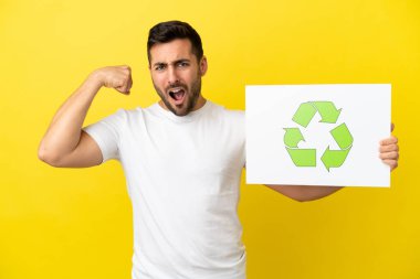 Sarı arka planda izole edilmiş yakışıklı beyaz adam elinde geri dönüşüm simgesi olan bir levha tutuyor ve güçlü hareketler yapıyor.
