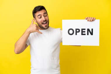 Sarı arka planda izole edilmiş yakışıklı beyaz bir adam. Elinde açık yazılı bir levha var ve telefon hareketi yapıyor.