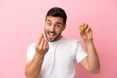 Elinde pembe arka planda izole edilmiş bir Bitcoin tutan genç adam para hareketi yapıyor.