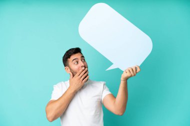 Mavi arka planda izole edilmiş yakışıklı beyaz bir adam elinde şaşırmış bir ifadeyle boş bir konuşma balonu tutuyor.