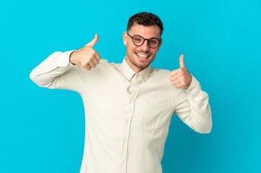 Beyaz tenli yakışıklı adam mavi arka planda izole edilmiş bir şekilde baş parmak işareti yapıyor.