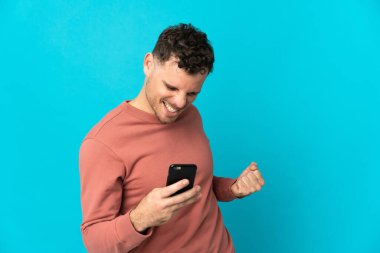 Mavi arka planda izole edilmiş genç beyaz adam cep telefonu kullanıyor ve zafer jesti yapıyor.