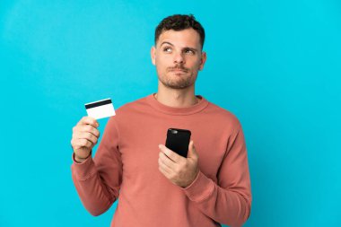 Kafkasyalı yakışıklı genç adam mavi arka planda izole edilmiş. Cep telefonuyla kredi kartıyla alışveriş yapıyor.