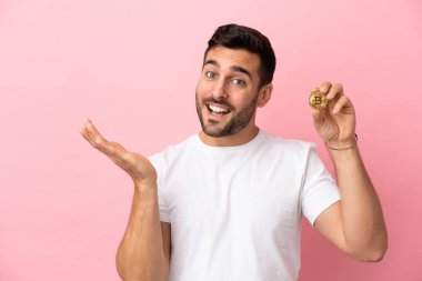 Pembe arka planda izole edilmiş Bitcoin 'i tutan genç adam davet etmek için ellerini yana uzatıyor.