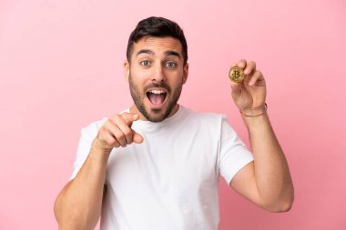 Elinde pembe arka planda izole edilmiş bir Bitcoin tutan genç adam şaşırmış ve ön tarafı işaret ediyor.