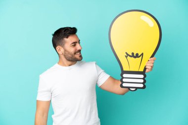 Mavi arka planda izole edilmiş yakışıklı beyaz bir adam elinde mutlu bir ifadeyle bir ampul simgesi tutuyor.