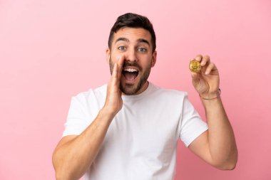 Elinde pembe arka planda izole edilmiş bir Bitcoin tutan genç adam ağzı açık bağırıyor.