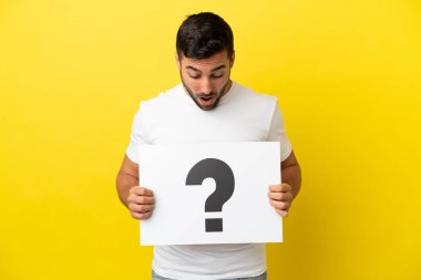 Sarı arka planda izole edilmiş, üzerinde soru işareti olan bir pankart taşıyan yakışıklı beyaz bir adam.