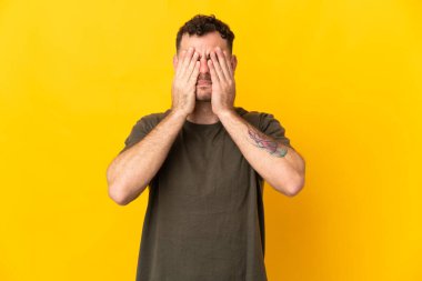 Beyaz tenli yakışıklı adam sarı arka planda izole edilmiş gözlerini elleriyle kapatıyor.