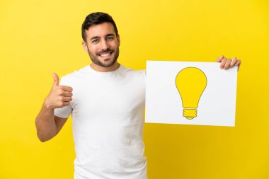 Sarı arka planda izole edilmiş yakışıklı beyaz bir adam elinde başparmağı havada bir ampul simgesi olan bir pankart tutuyor.