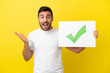 Sarı arka planda izole edilmiş yakışıklı beyaz bir adam. Elinde bir pankart var. Üzerinde yeşil işaret simgesi ve şaşırmış ifade var.