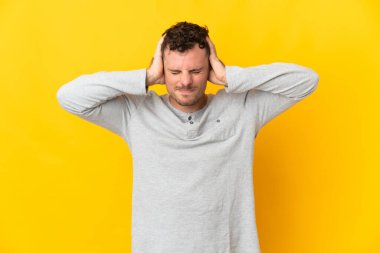 Sarı arka planda izole edilmiş genç beyaz yakışıklı adam hayal kırıklığına uğramış ve kulaklarını kapatmış.
