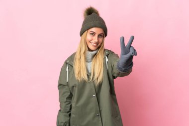 Kış şapkalı genç kadın pembe arka planda izole edilmiş gülümsüyor ve zafer işareti gösteriyor.