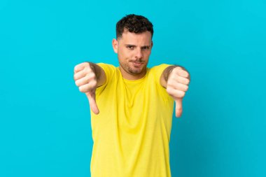 Mavi arka planda izole edilmiş, iki eliyle baş parmağını gösteren beyaz tenli yakışıklı bir adam.