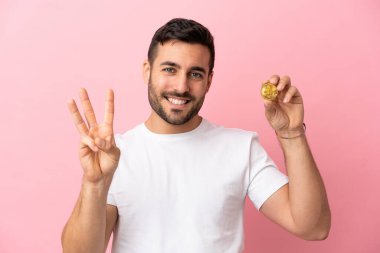 Elinde pembe arka planda izole edilmiş Bitcoin tutan genç adam mutlu ve parmaklarıyla üçe kadar sayıyor.