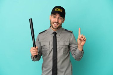 Genç güvenlik elemanı mavi arka planda izole edilmiş ve en iyinin işareti olarak parmağını kaldırıyor.