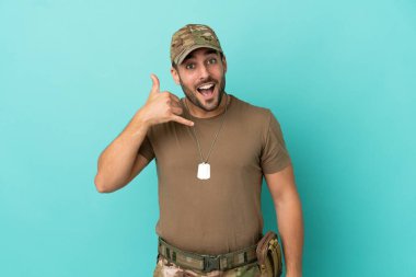 Mavi arka planda izole edilmiş künyeli bir ordu telefon jesti yapıyor. Bana geri dönüş işareti ver.