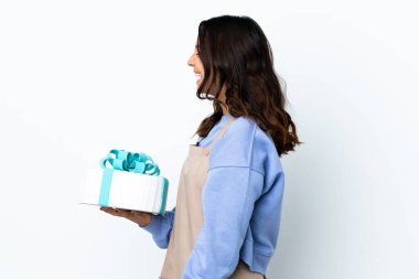 Pasta şefi, beyaz arka planda büyük bir pasta tutuyor. Yan pozisyonda gülüyor.