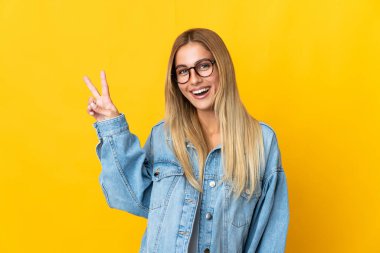 Sarı arka planda izole edilmiş genç sarışın kadın gülümsüyor ve zafer işareti gösteriyor.
