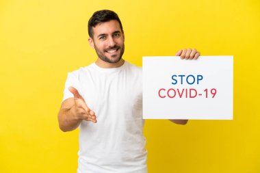 Sarı arka planda izole edilmiş yakışıklı beyaz bir adam elinde yazılı bir levhayla Covid 19 'u durdur ve bir anlaşma yap.