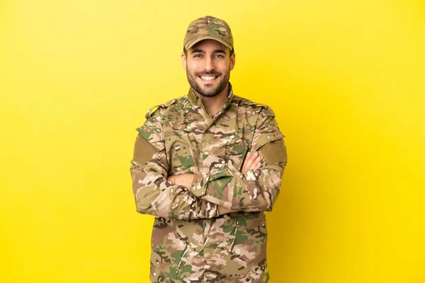 Military man isolated on yellow background keeping the arms crossed in frontal position