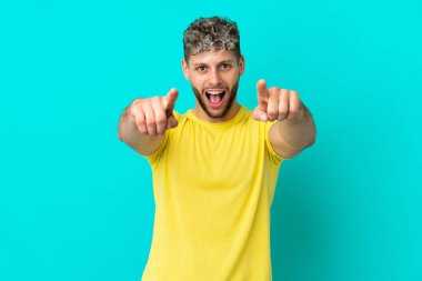 Mavi arka planda izole edilmiş yakışıklı beyaz adam şaşırmış ve ön tarafı işaret ediyor.