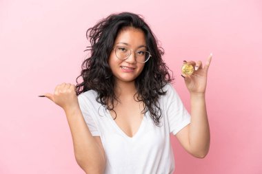 Pembe arka planda Bitcoin tutan Asyalı genç bir kadın bir ürünü sunmak için yan tarafı işaret ediyor.