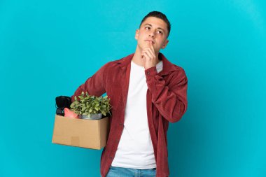 Kafkasyalı genç adam mavi arka planda izole edilmiş eşyalarla dolu bir kutuyu kaldırırken hareket ediyor.