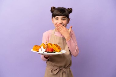 Mor arka planda izole edilmiş waffle tutan küçük beyaz kız mutlu ve ağzı eliyle kaplayan gülümsüyor.