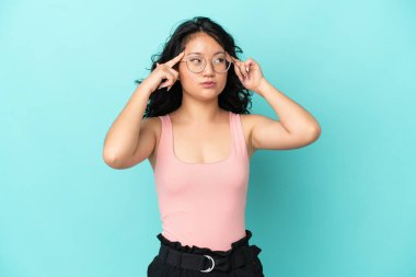 Asyalı genç bir kadın mavi arka planda izole edilmiş. Şüpheleri ve düşünceleri var.