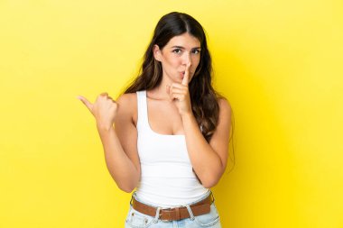 Sarı arka planda izole edilmiş genç, beyaz bir kadın. Yan tarafı işaret ediyor ve sessizlik hareketi yapıyor.