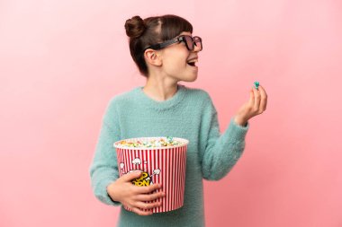 Küçük beyaz kız pembe arka planda 3 boyutlu gözlüklerle izole edilmiş ve elinde koca bir kova patlamış mısır tutuyor.