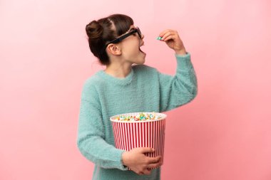 Küçük beyaz kız pembe arka planda 3 boyutlu gözlüklerle izole edilmiş ve elinde koca bir kova patlamış mısır tutuyor.