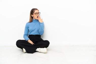 Beyaz bir kadın yerde oturuyor esniyor ve ağzı açık bir şekilde elleriyle ağzını kapatıyor.