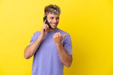 Sarı arka planda izole edilmiş cep telefonu kullanan genç beyaz adam bir ürünü sunmak için yan tarafı işaret ediyor.