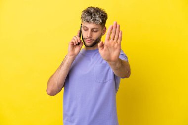 Sarı arka planda izole edilmiş cep telefonu kullanan genç beyaz adam dur hareketi yapıyor ve hayal kırıklığına uğruyor.