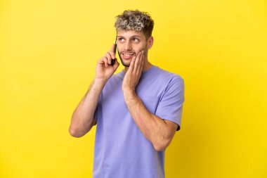 Sarı arka planda izole edilmiş cep telefonu kullanan beyaz bir adam gülümserken yukarı bakıyor.