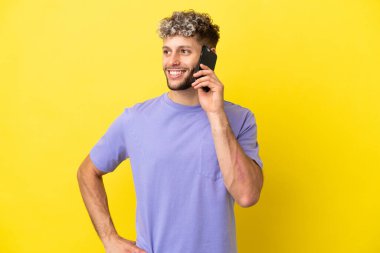 Sarı arka planda izole edilmiş cep telefonu kullanan beyaz bir adam. Kolları kalçasında ve gülümsüyor.