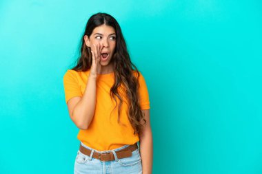 Mavi arka planda izole edilmiş genç beyaz kadın yan tarafa bakarken sürpriz bir jest yaparak bir şeyler fısıldıyor.