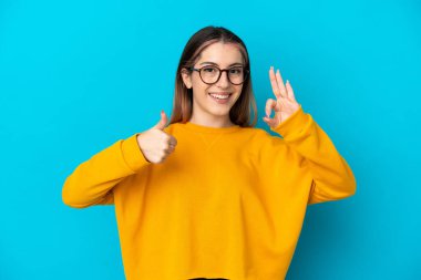 Mavi arka planda izole edilmiş genç beyaz kadın işaret ve baş parmak hareketi gösteriyor.